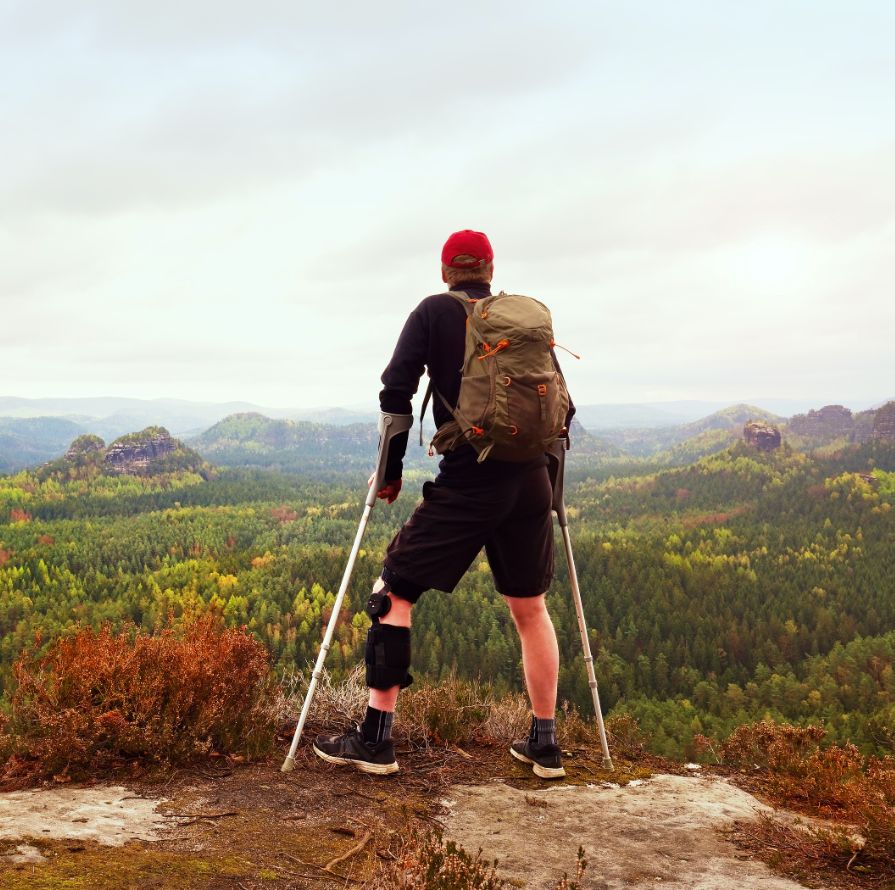 A Man With Crutches And A Leg Brace Stares Out At A Mountain Valley.