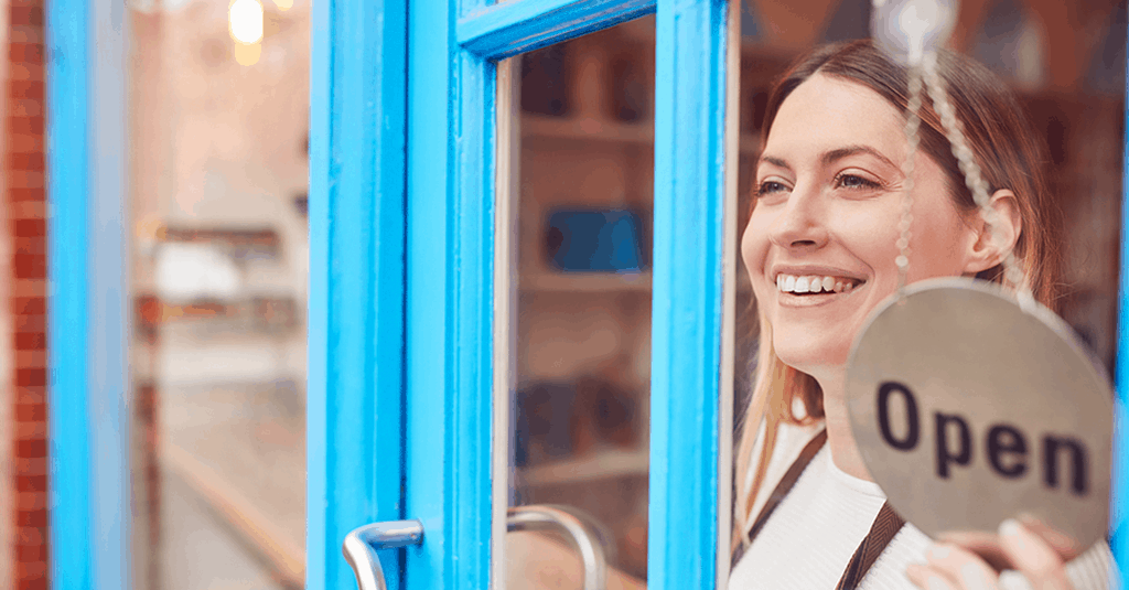 Smiling Woman Looking Out Of A Storefront Widow While Flipping Over An Open Sign
